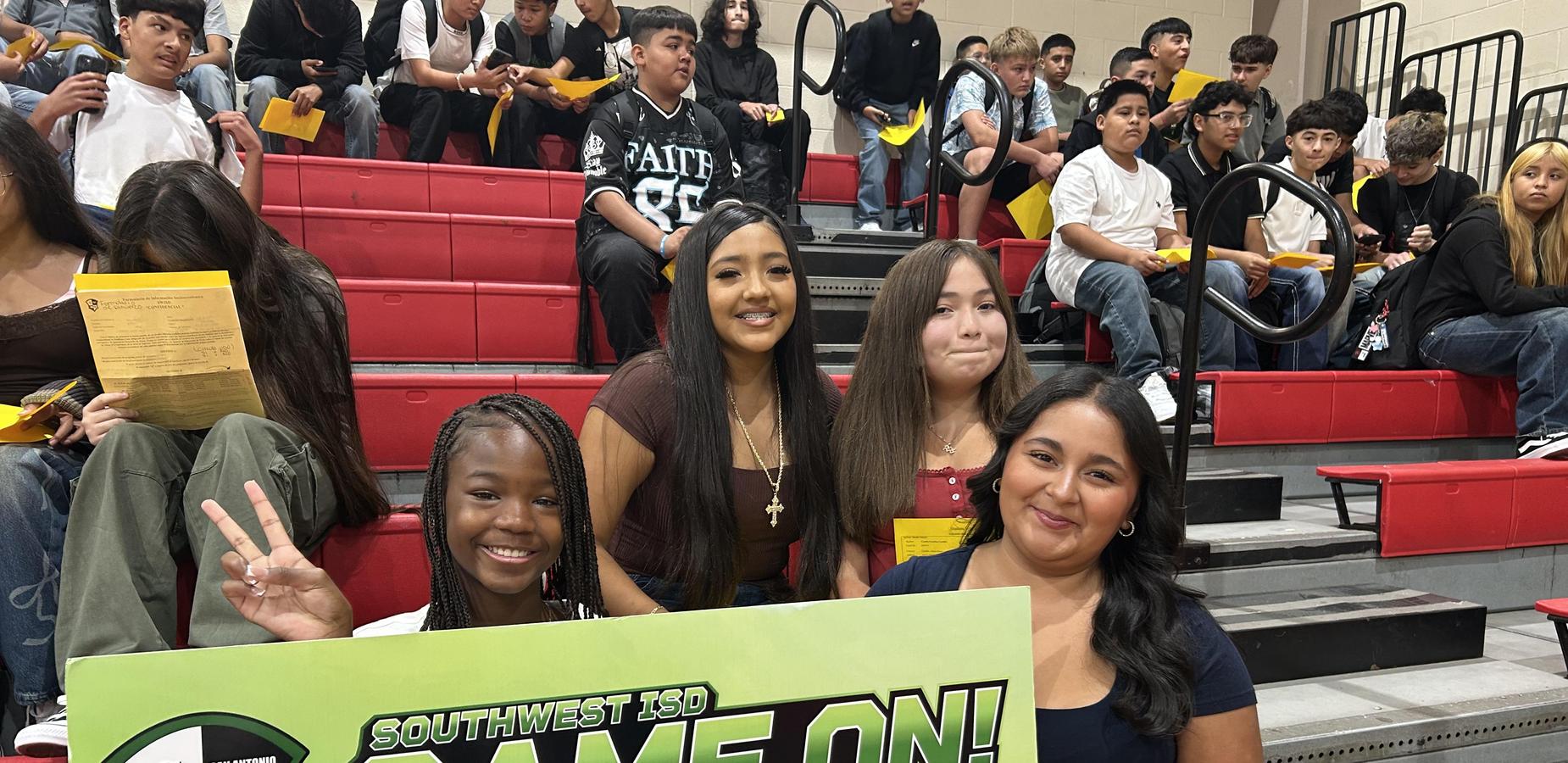 Four girls cheer in the bleachers, holding a green banner that reads 'GAME ON!'.