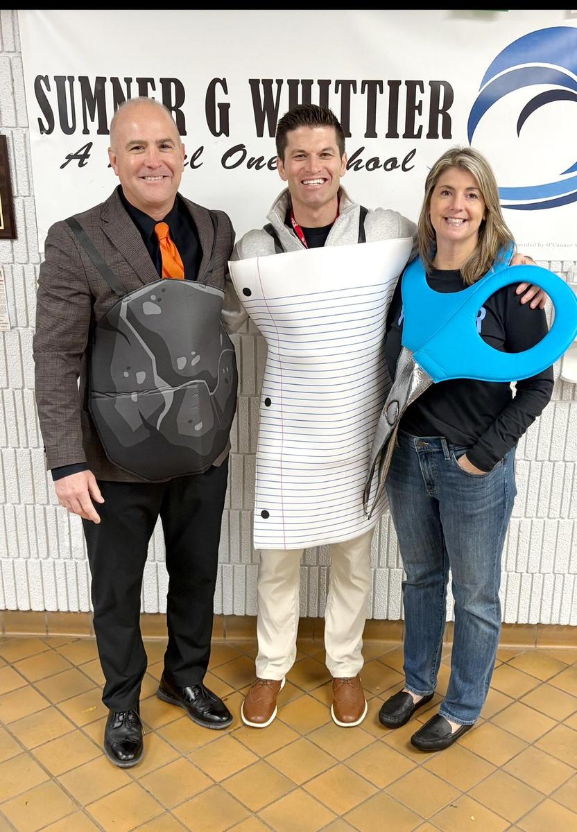 Three adults dressed as a pencil, scissors, and a paper sheet, smiling in a school hallway.