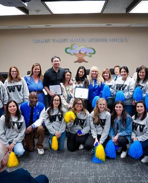 Group photo of cheerleaders holding certificates and props in a school district meeting.