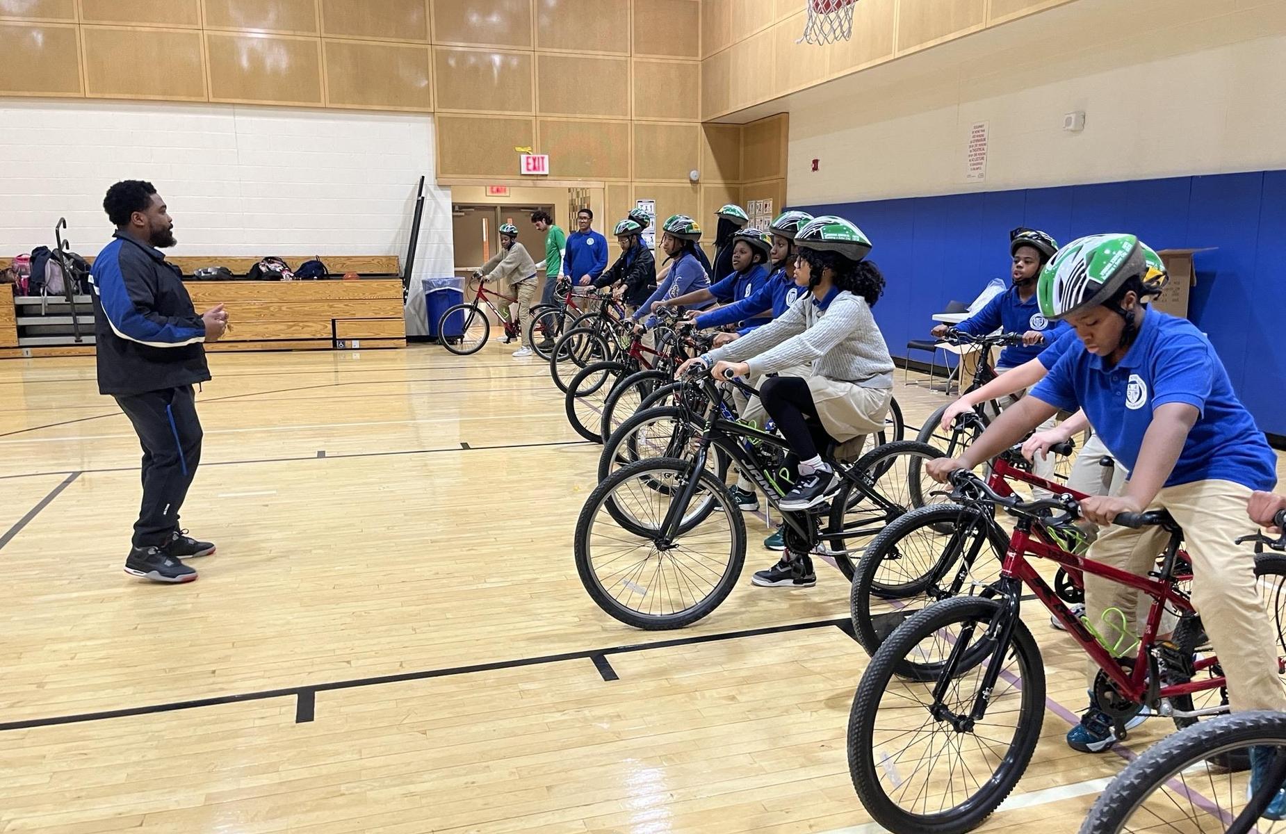 A coach instructs a group of youth on bicycles in a gymnasium.