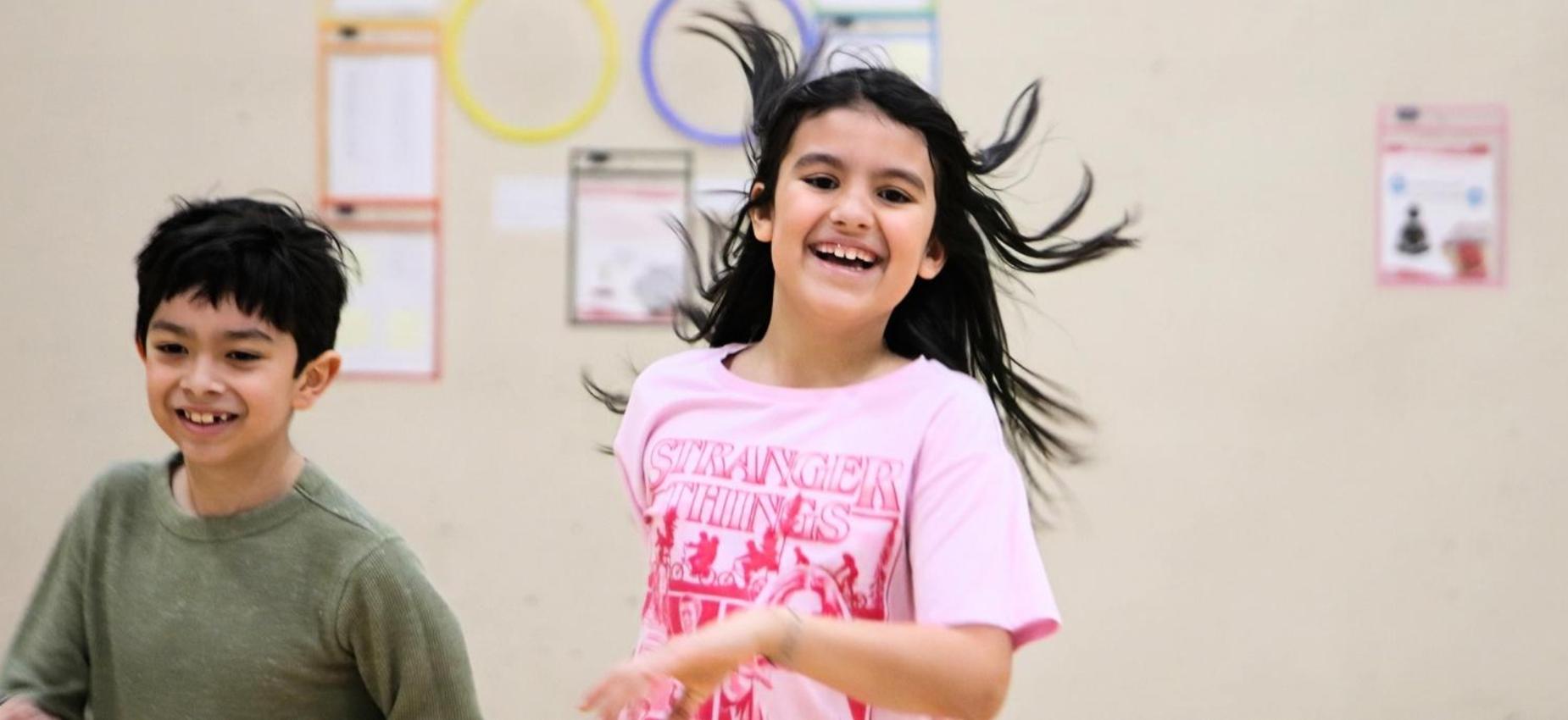Children play and run joyfully in a gym with colorful hoops in the background.