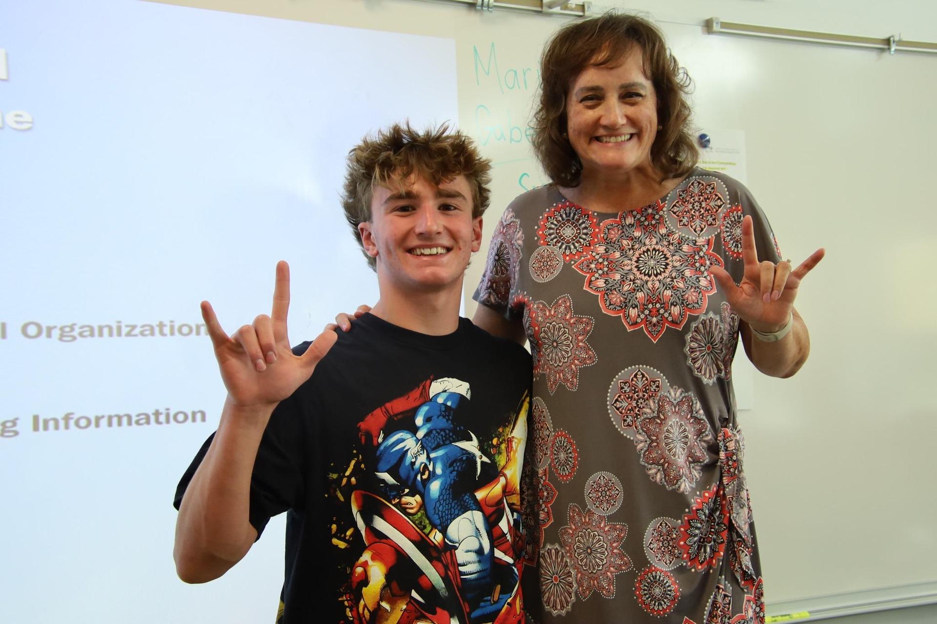 high school student and teacher holding up 'i love you' sign and smiling for camera in ASL classroom