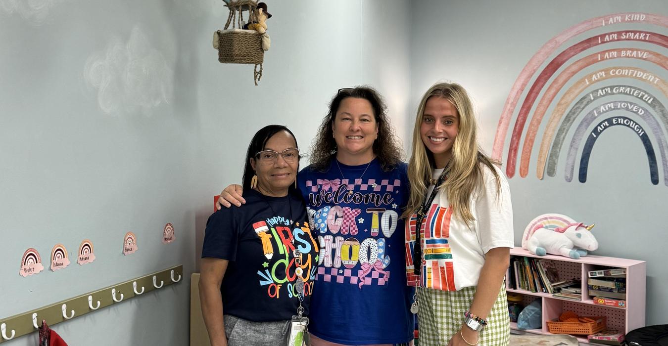 Three women stand together in a colorful classroom with a positive message wall.