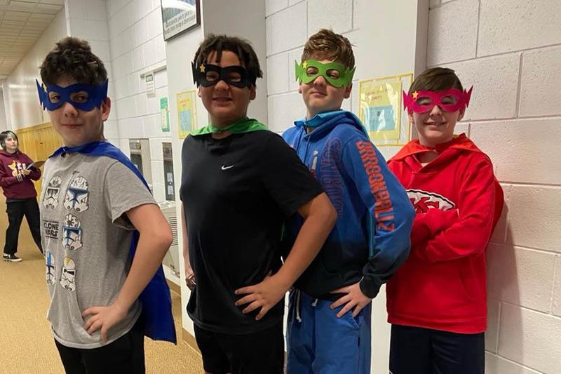 4 male students wear superhero masks and capes and stand with hands on hips