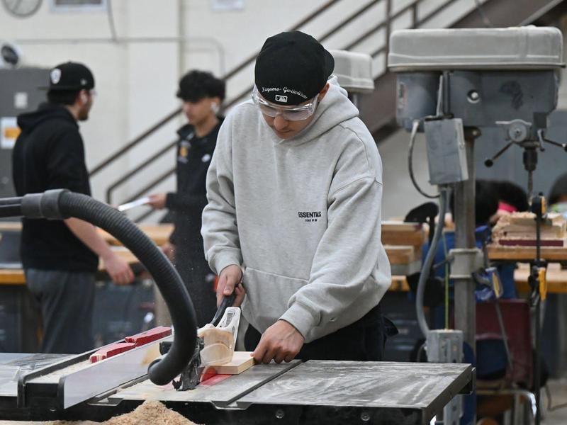 A student operating a table saw in a workshop filled with classmates.