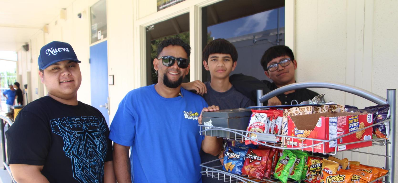 Four individuals pose next to a cart filled with various snacks and drinks.