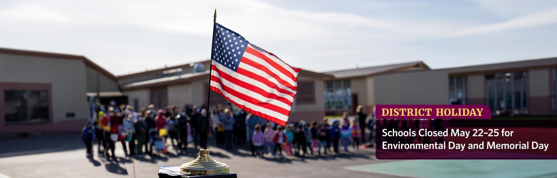 A small U.S. flag on a brass stand with students and families in the background on a school playground.