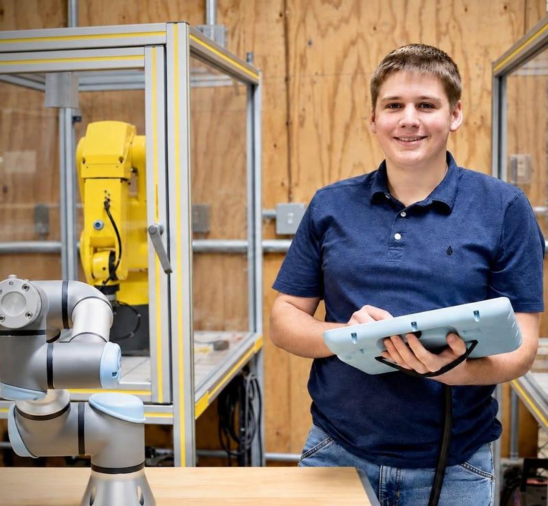 Male student, smiling, standing next to a robotic arm holding a controller