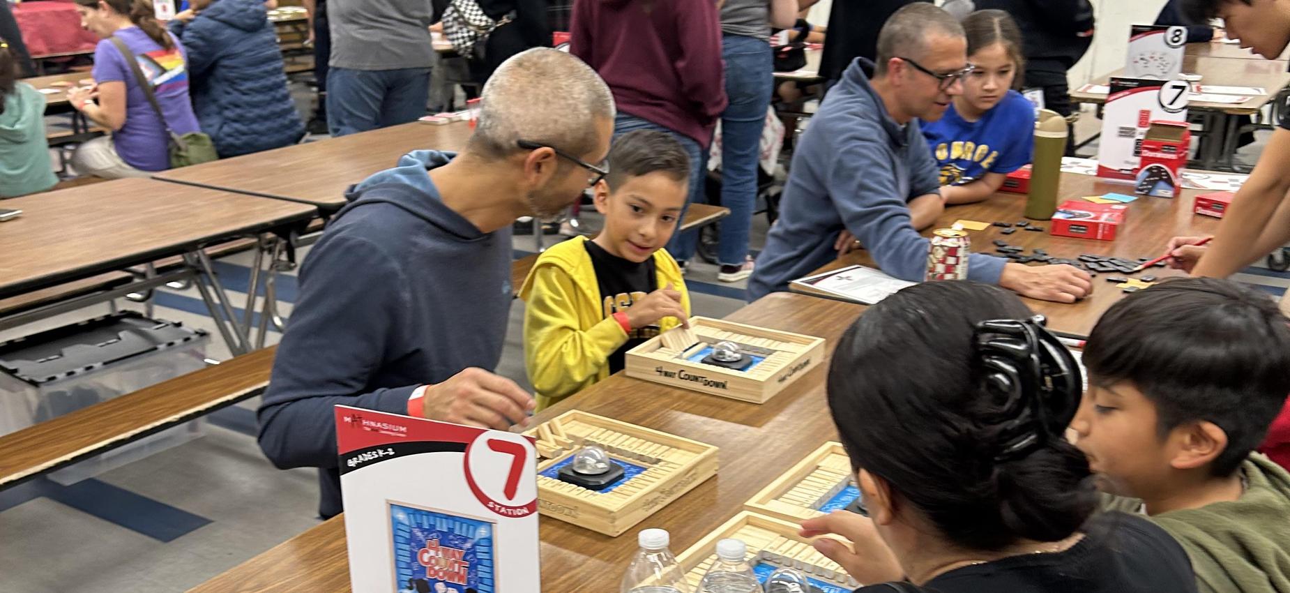Children and adults are gathered around tables playing board games together.