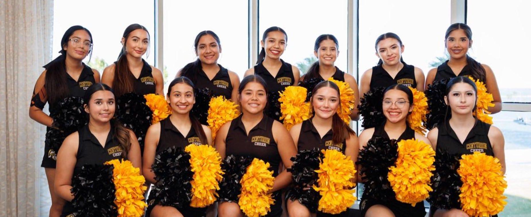 A group of cheerleaders posing in front of large windows, holding pom-poms.