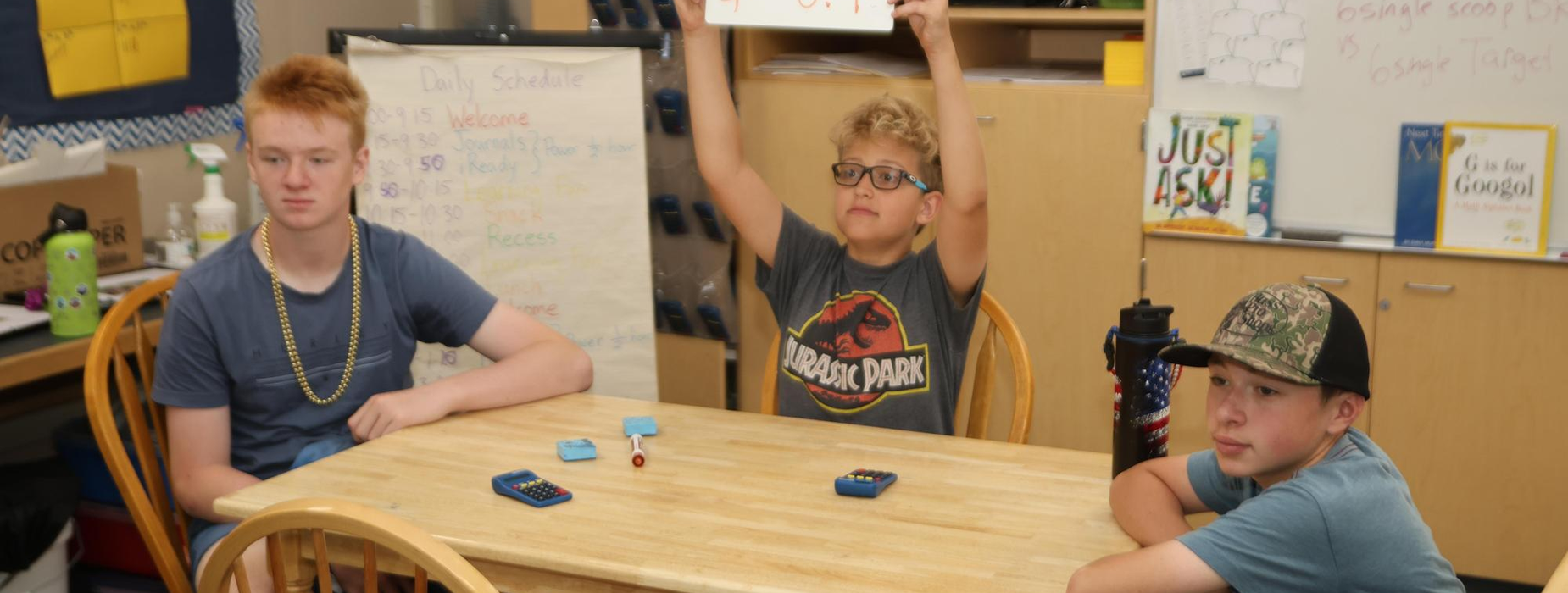 Three boys sitting at a table, one holding up a sign while others look on.