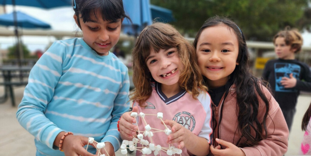 Three girls smile while building a structure with marshmallows and sticks.