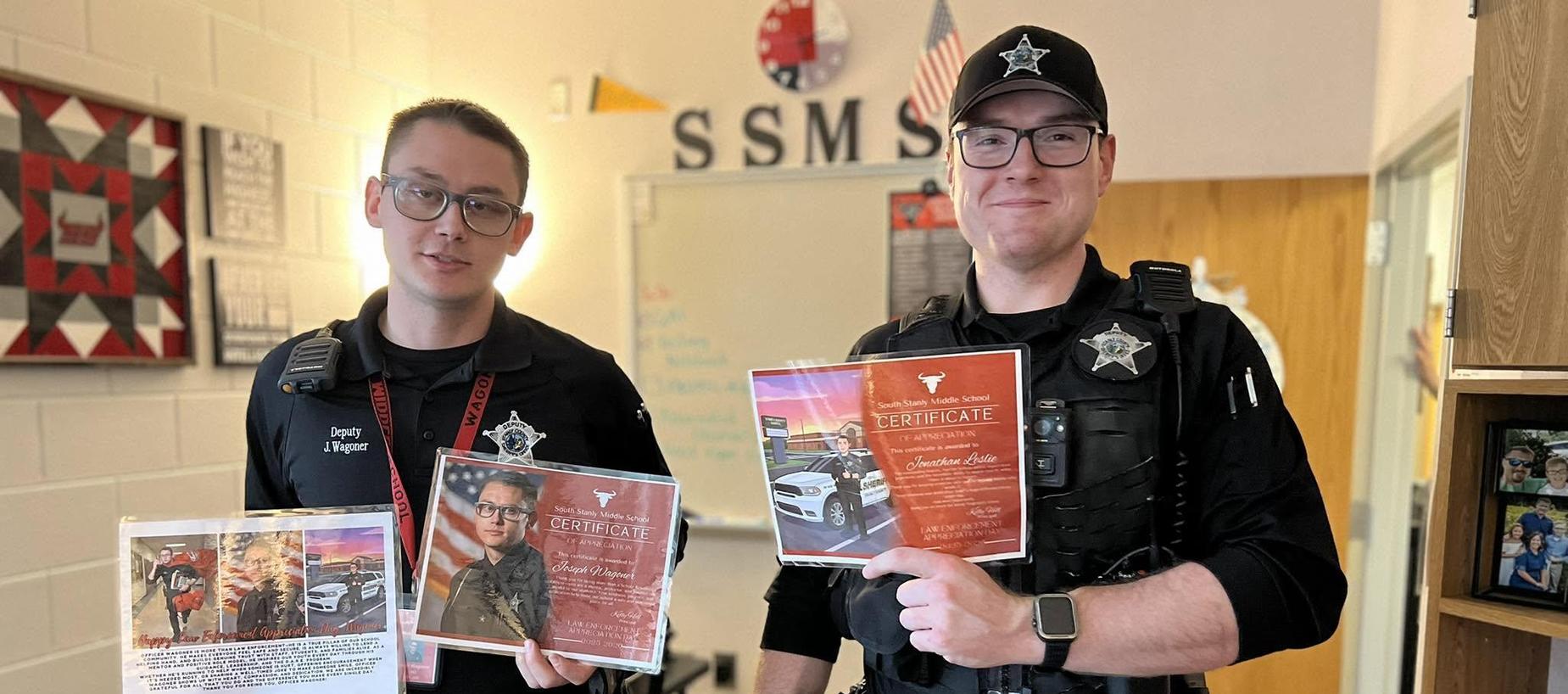 Two police officers hold certificates and awards in a classroom setting.