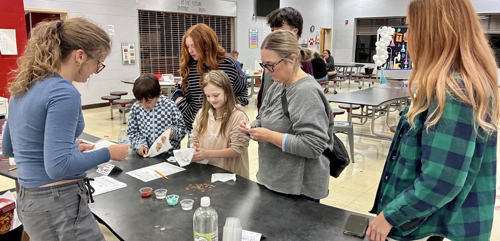 Students and adults gathered around a table with plastic bottles and small sample cups for a science experiment