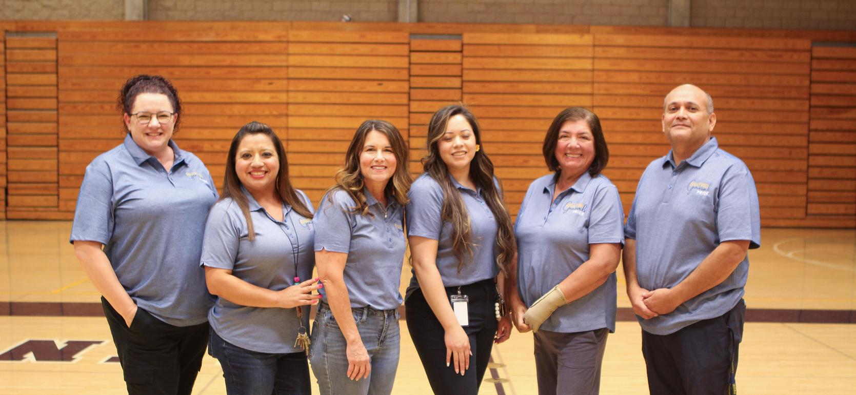 Six staff members in blue shirts pose for a group photo in a gymnasium.