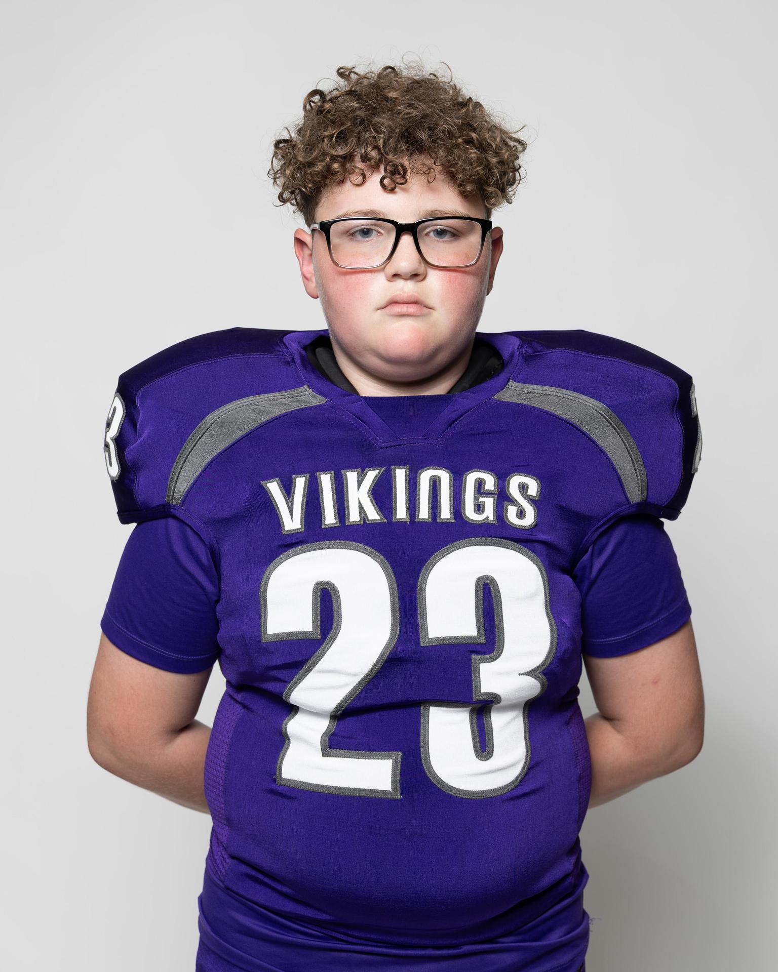 Boy with curly hair in glasses wearing a purple Vikings jersey, arms crossed.