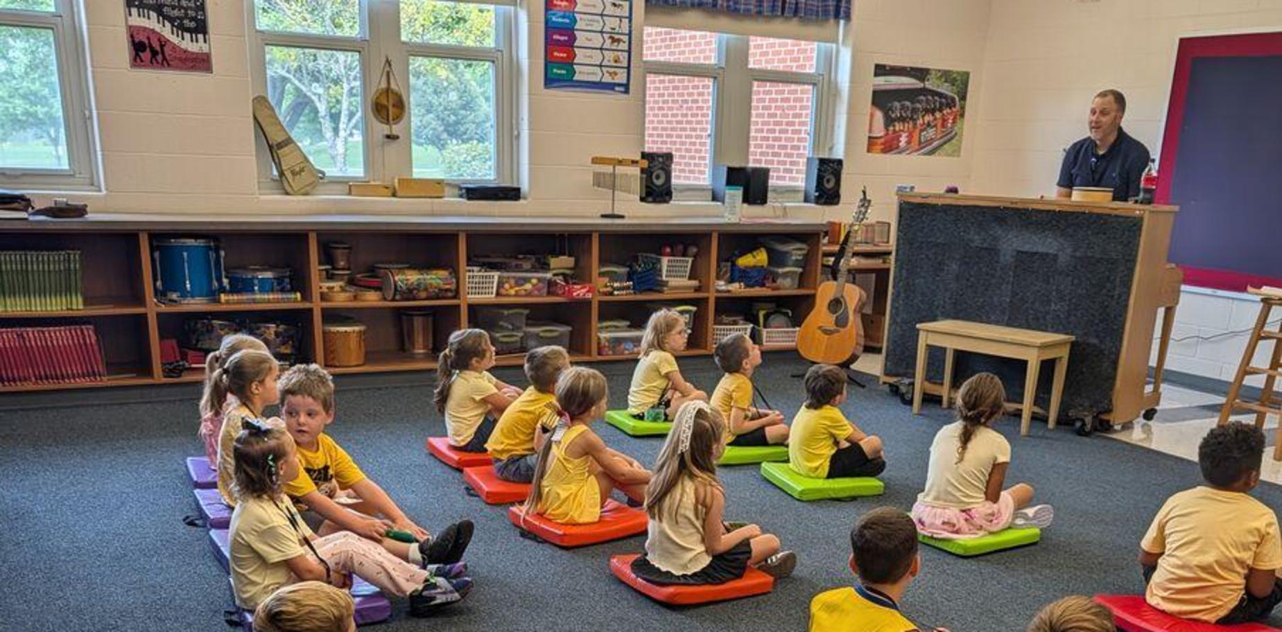 Young children sit on colorful mats, attentively listening in a classroom.