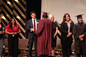 A line of dignitaries and faculty, including administrators and educators, congratulates a graduate in a maroon cap and gown during the ceremony on stage.