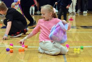 Child in a pink sweater collecting colorful eggs on a gym floor.