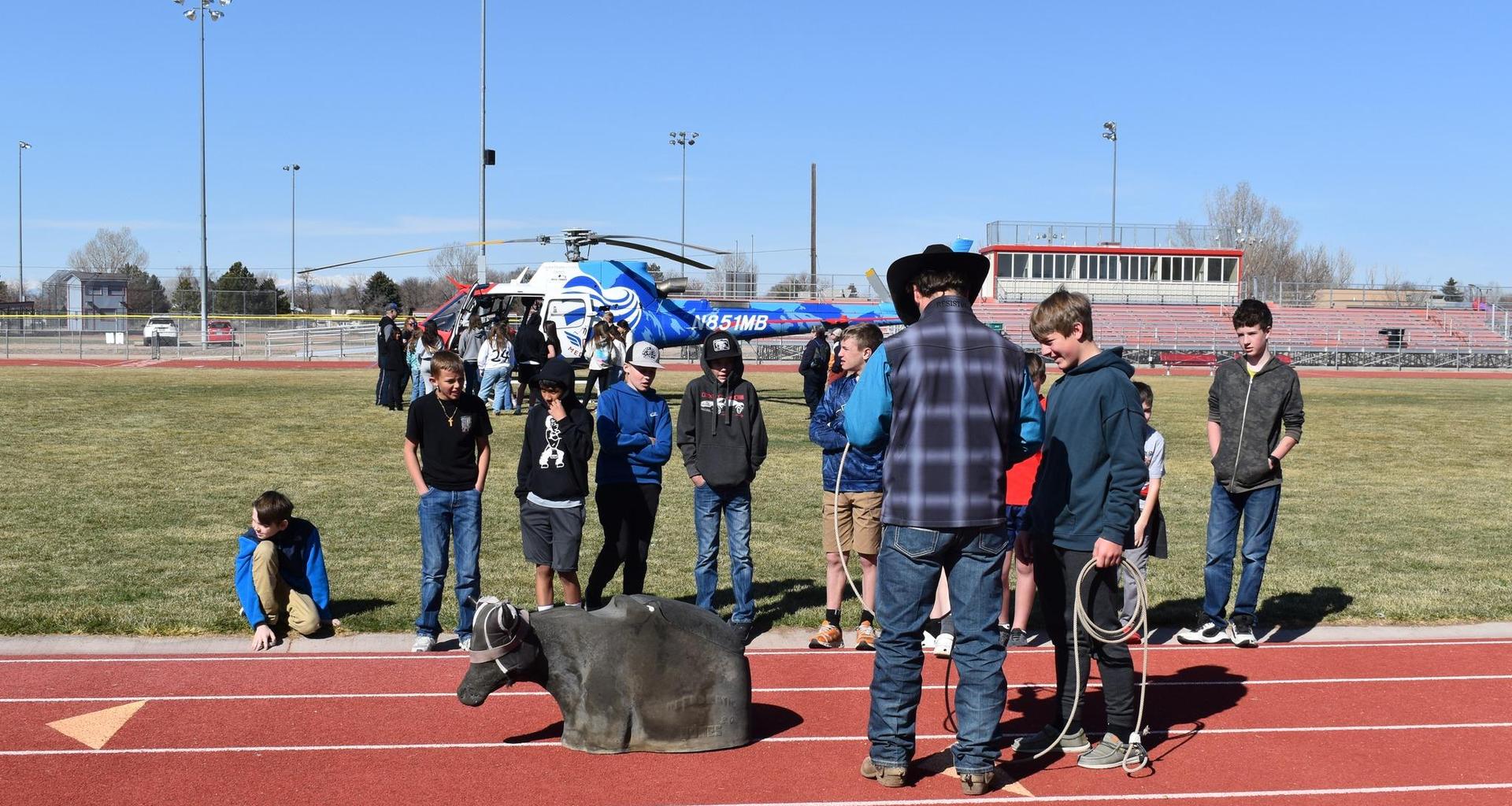 A group of children watches a cowboy demonstrating lasso techniques with a practice cow on a track.