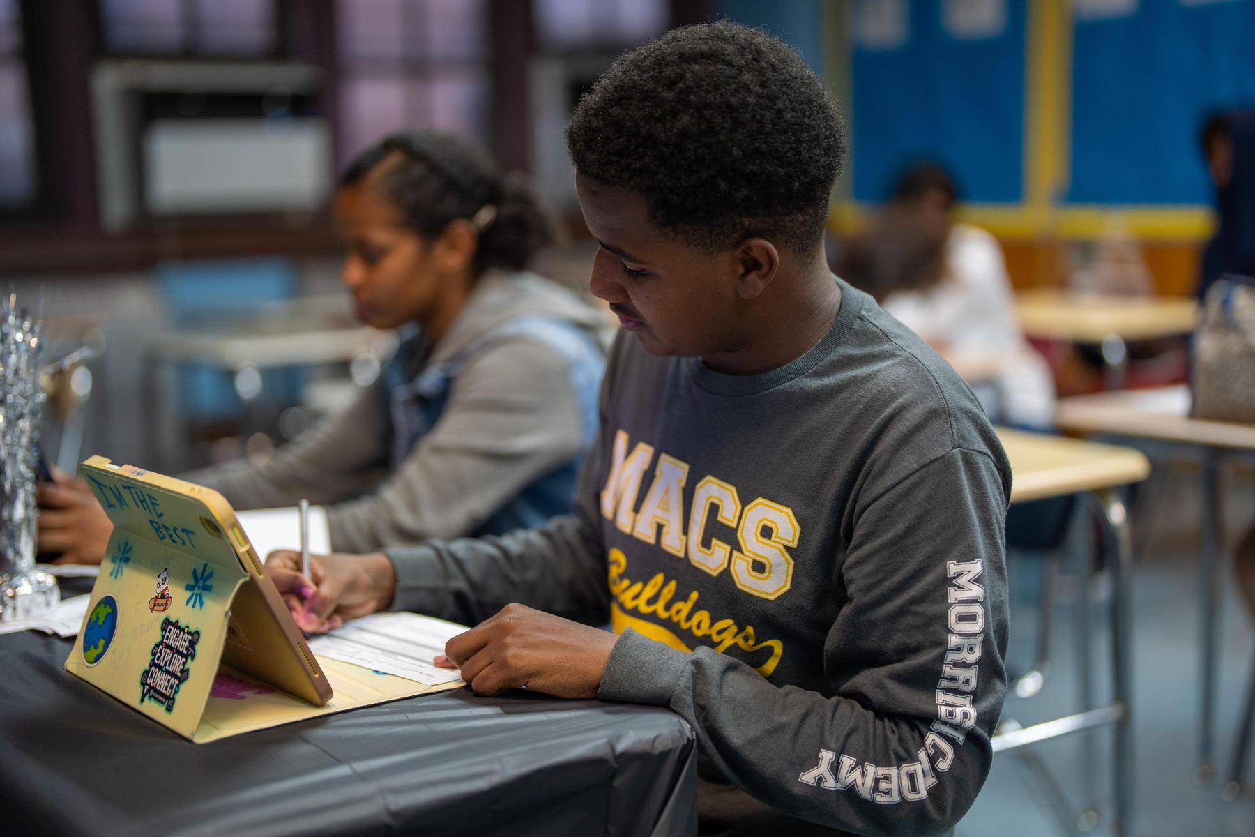 A student writing at a desk in a classroom with peers in the background.
