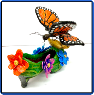 Paper mache monarch butterfly resting on the tip of a green shoe with colorful flowers