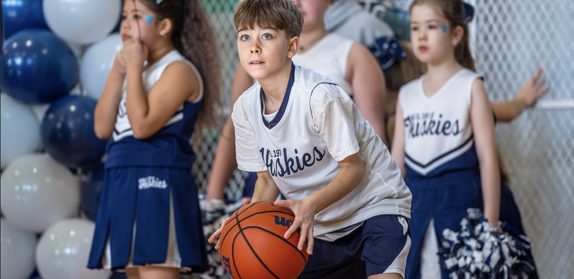 A boy in a basketball uniform holds a ball, with cheerleaders in the background.