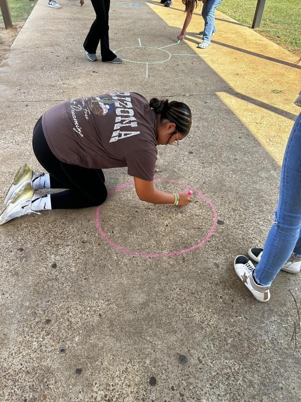 Student writing on concrete outside with pink chalk