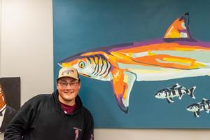 Student in front of his artwork, a large painting of a shark.