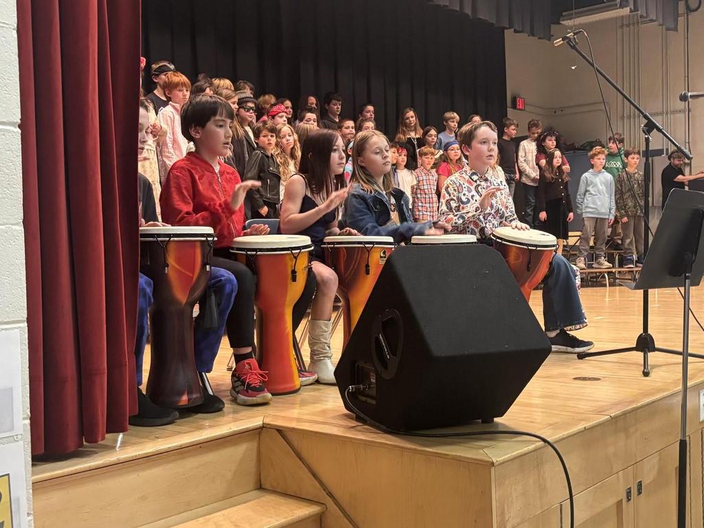 Elementary students on stage playing bongo drums and on bleachers singing.
