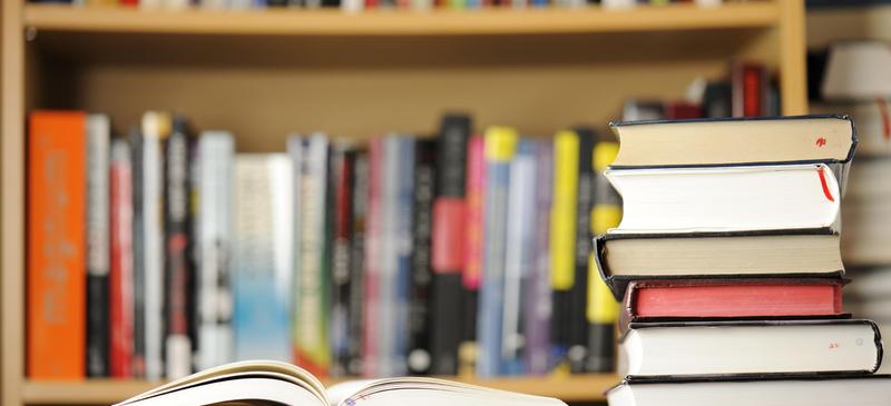 Opened book in front of a stack of books on a shelf filled with various titles.