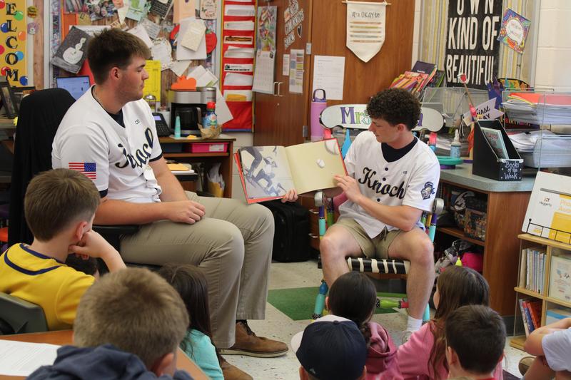 2 baseball players sit in front of classroom reading a book