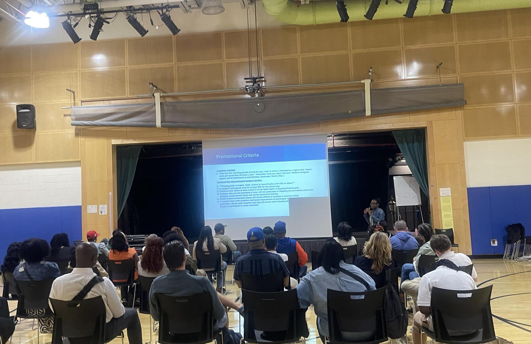Audience seated in a gymnasium watching a presentation on a screen.