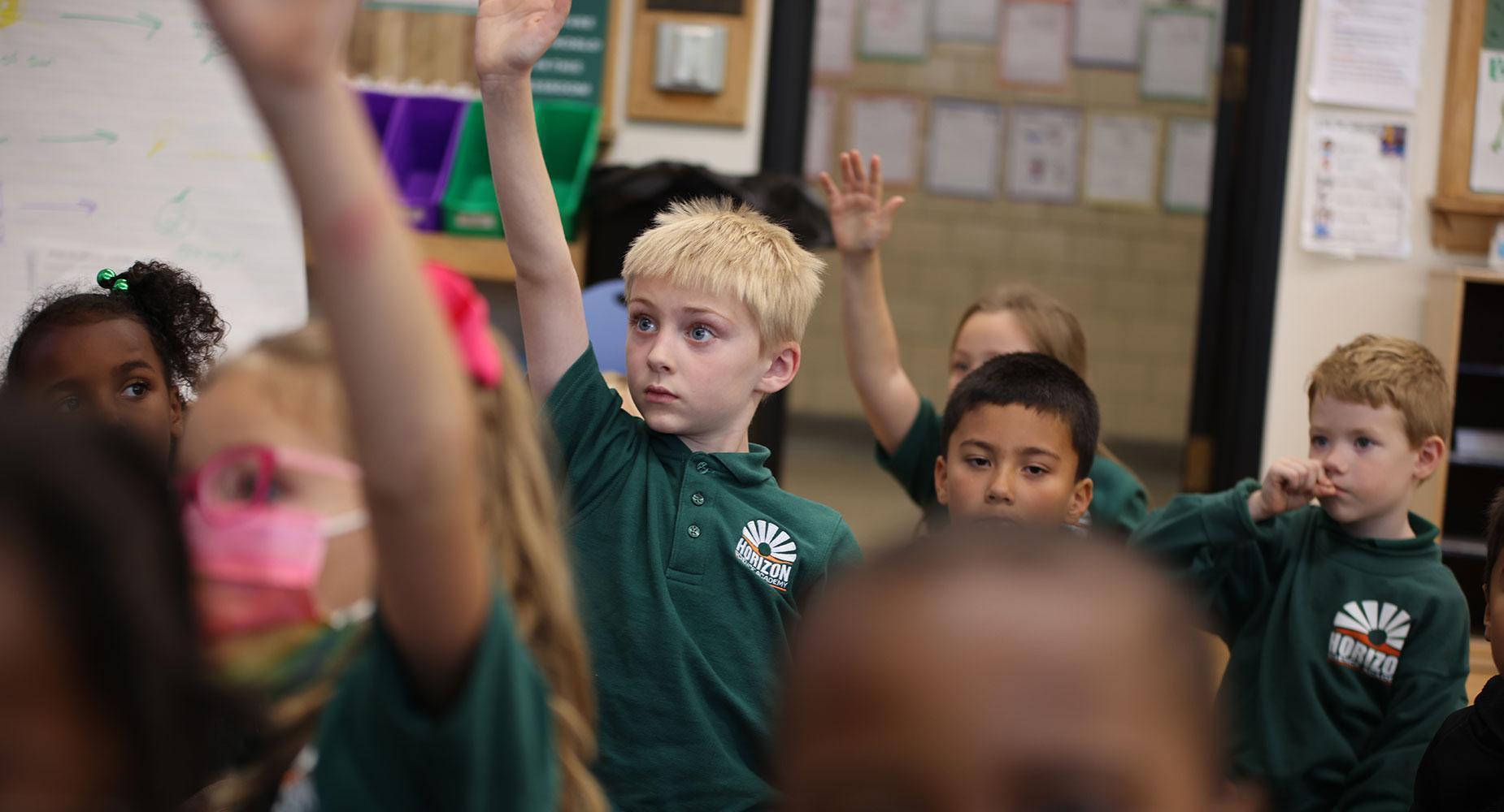 Students raising hands in a classroom with a focus on one boy.