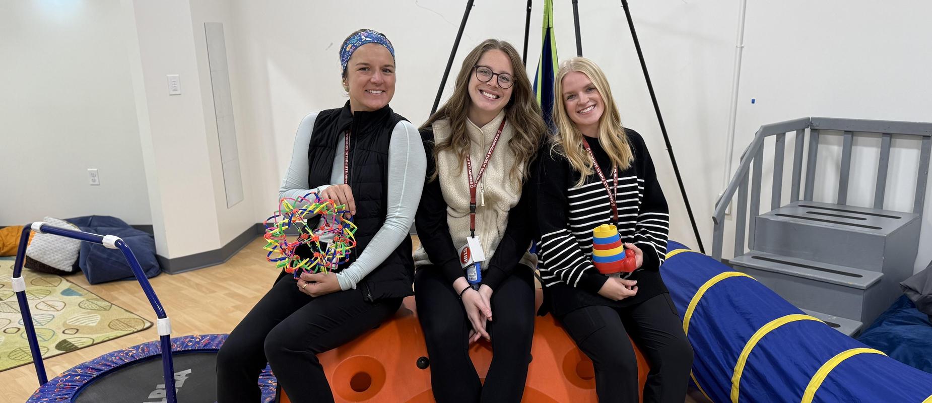 three occupational therapists smile in their OT room with their various gadgets for therapy