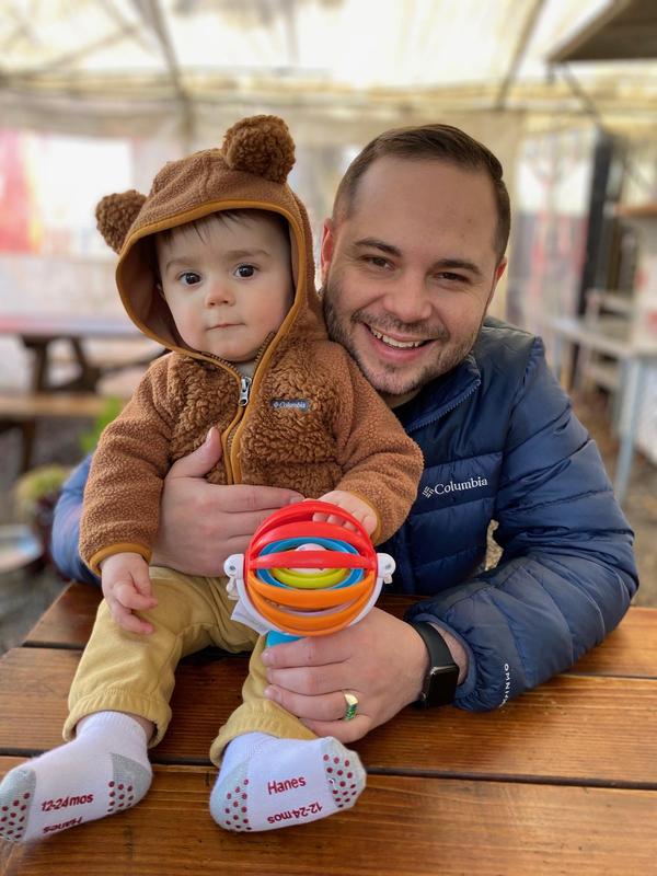 David Zahn, PE teacher for Arco Iris Spanish Immersion School, sits at a wooden table hugging his baby son who sits on top of it.