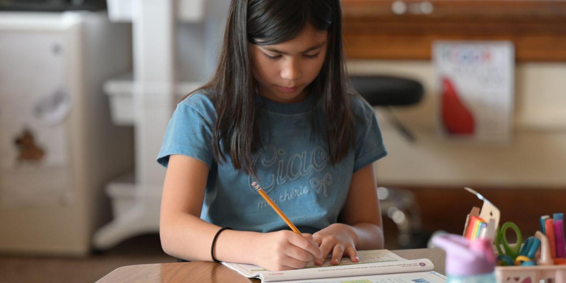 A girl writing in a book at a classroom table.