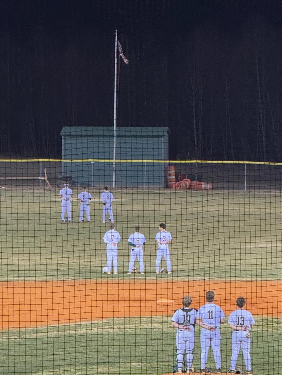 Baseball players lined up in front of a flagpole during a nighttime game.