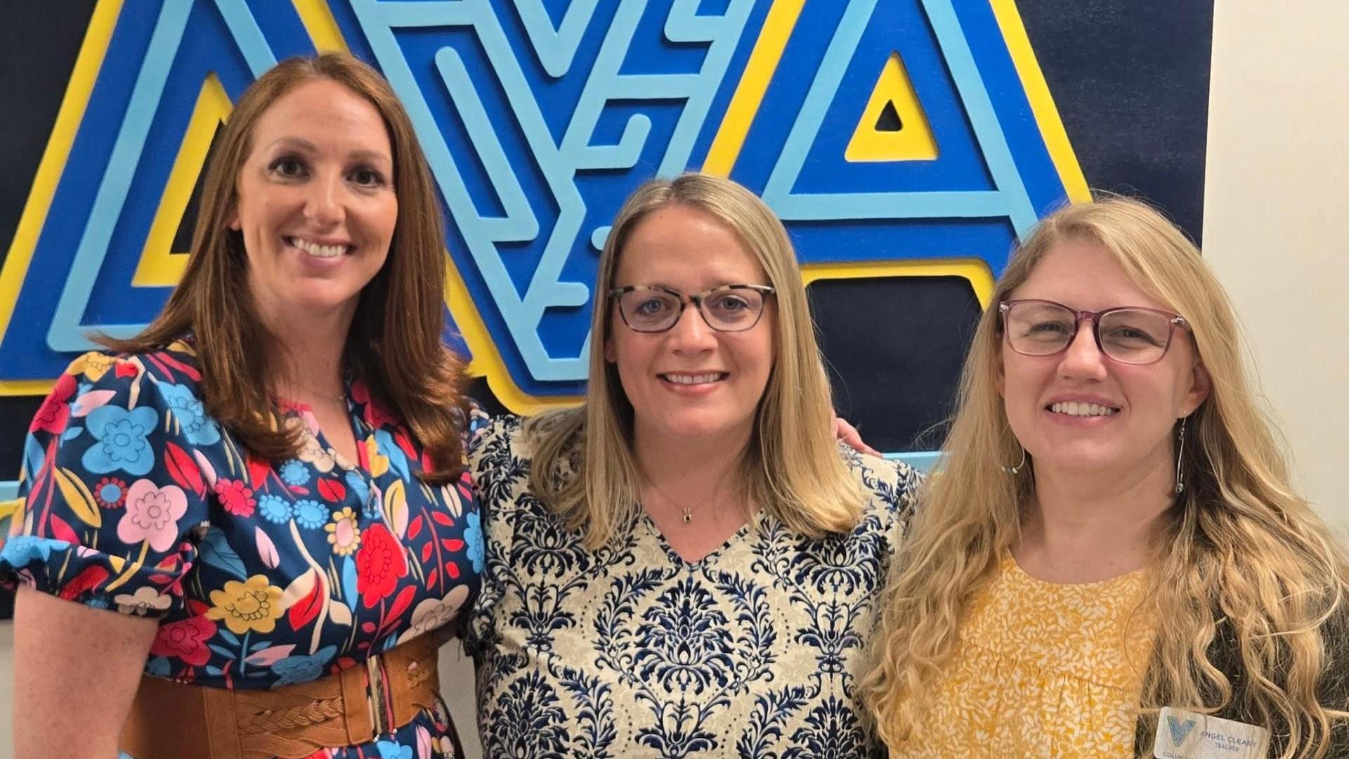 Three women pose together in front of a colorful blue and yellow logo.