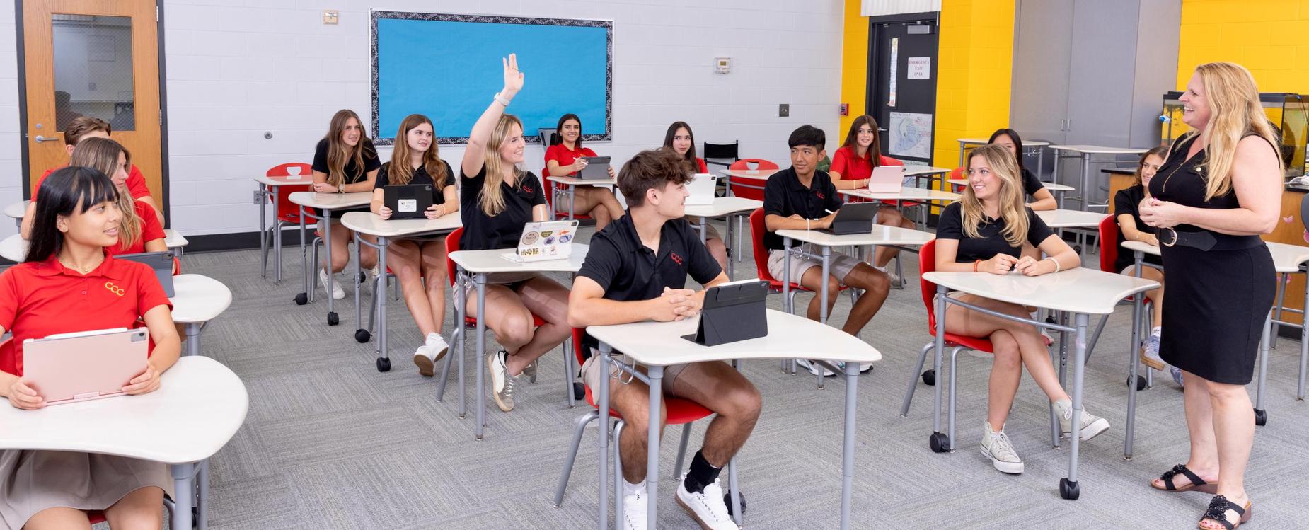 Classroom full of students in red and black polos seated at desks lined up in rows. One girl in the middle of the classroom raises her hand as she and teacher at front of room smile at each other.
