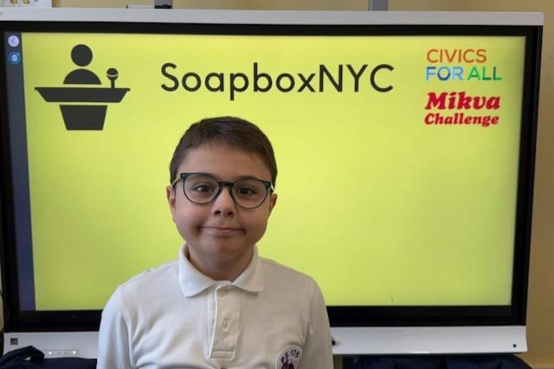 Child in glasses stands in front of a bright yellow screen displaying 'SoapboxNYC' and logos.