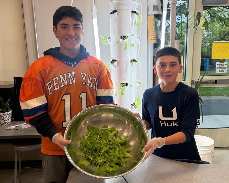 Two students hold a large metal bowl filled with harvested lettuce in front of an indoor Grow Tower.