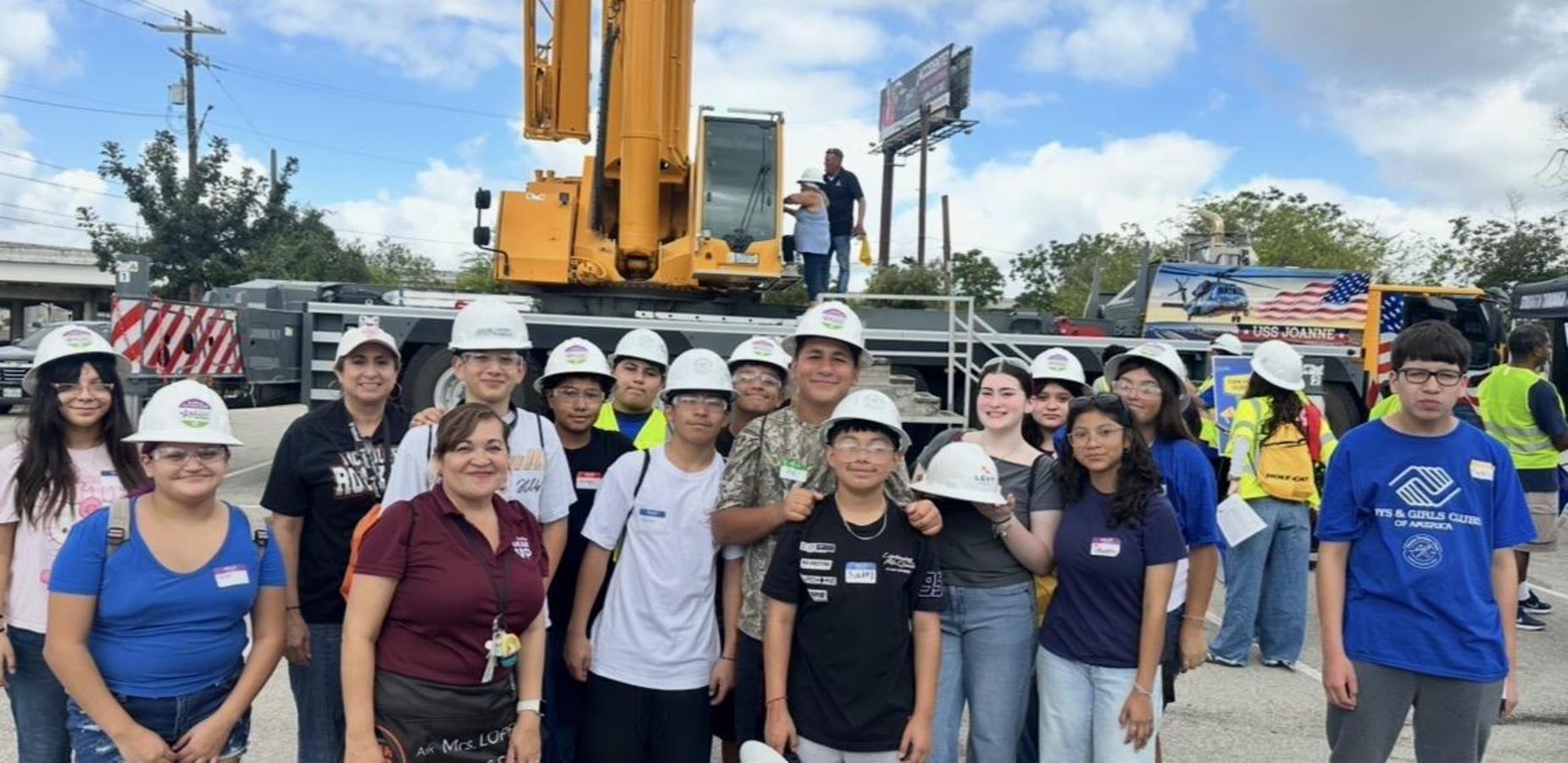 Group of students and adults posing with construction equipment at an outdoor event.