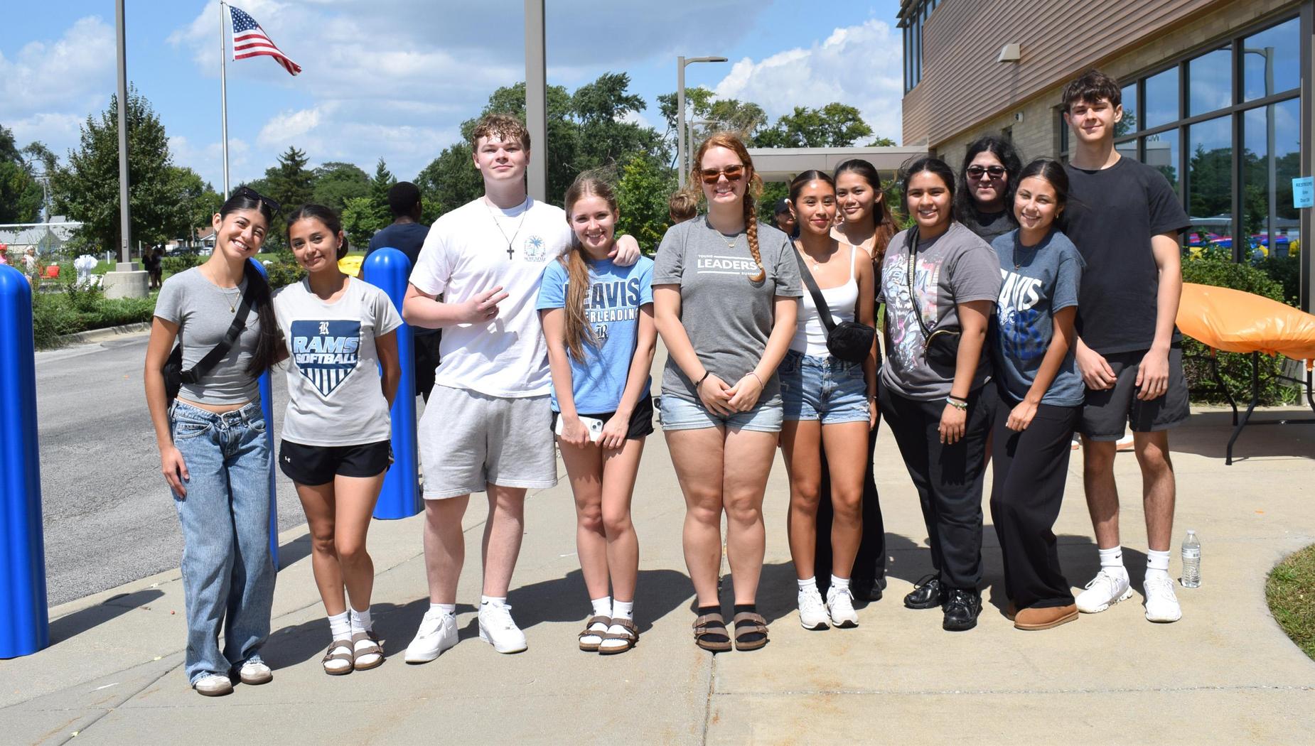 Group of twelve young people posing outside a building with an American flag.
