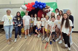 Group of children and adults smiling together in front of a colorful balloon arch.