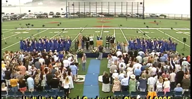 Graduation ceremony with graduates in blue gowns, audience, and a decorated stage.