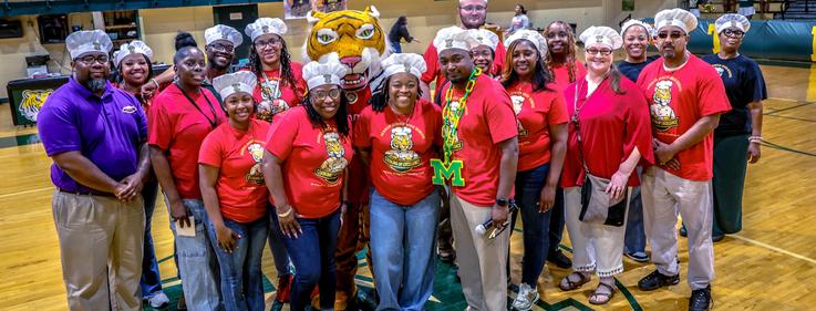 Group of people in chef hats posing together in a gymnasium.