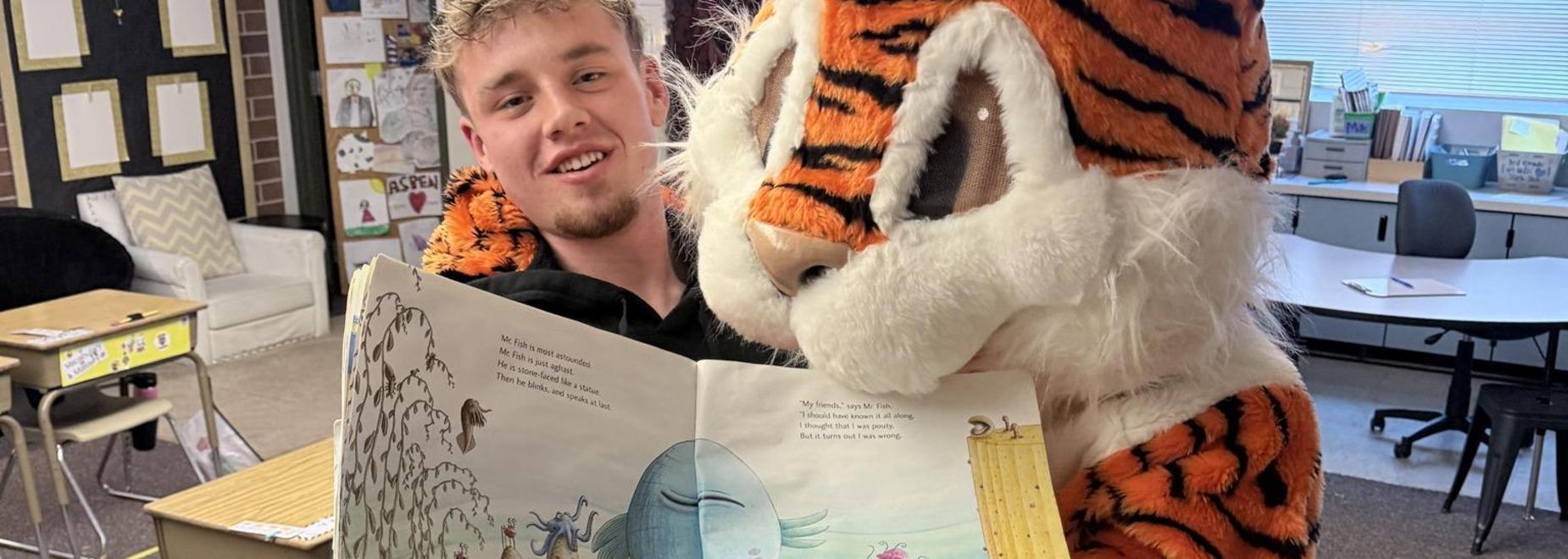 A person in a tiger costume reads a storybook to a student.