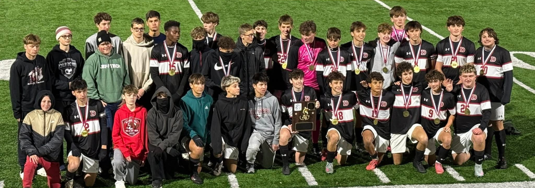 Group photo of the winning boys' soccer team displaying medals and a trophy on a field.