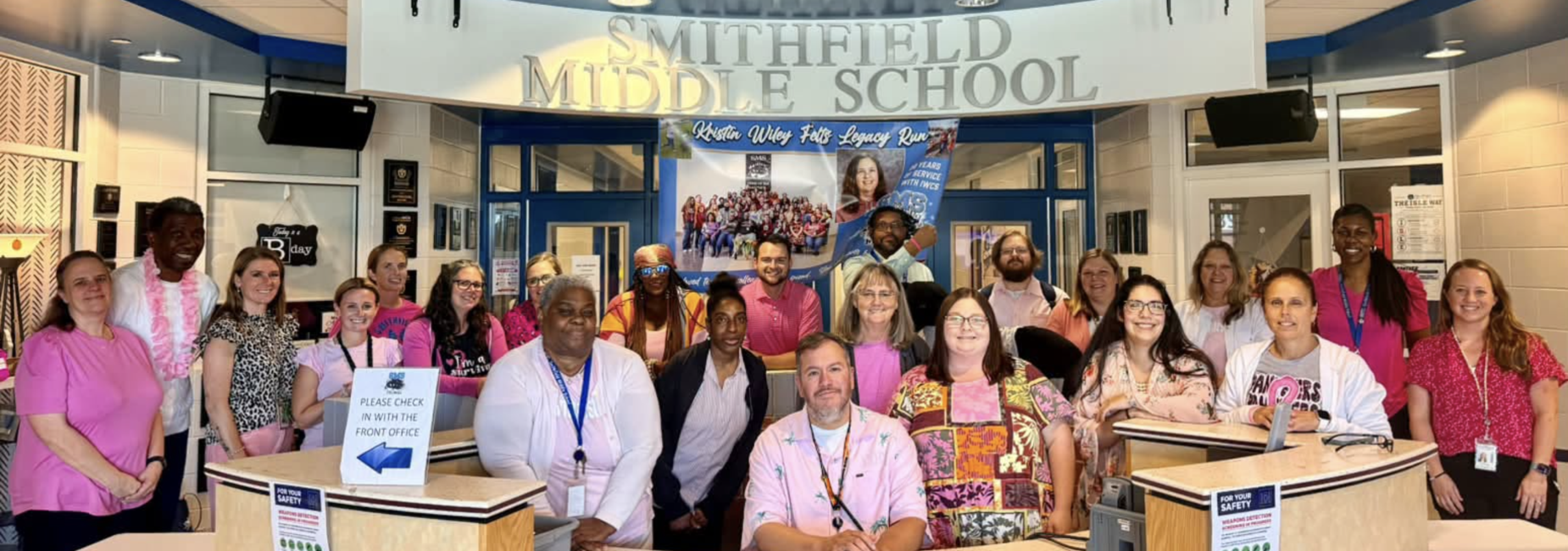 Group of staff members at Smithfield Middle School wearing pink, smiling in a lobby.